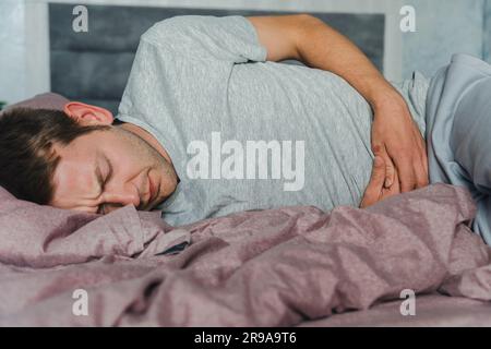 Frustrated handsome young man hugging his belly suffering from stomach pain while lying in bad at home. Male with belly ache. Stomach ulcer, gastritis Stock Photo