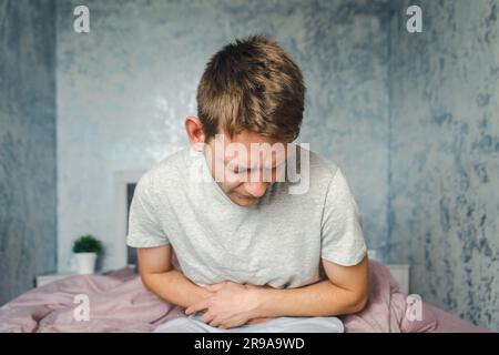 Frustrated handsome young man hugging his belly suffering from stomach pain with closed eyes while sitting on the couch at home. Male with belly ache Stock Photo