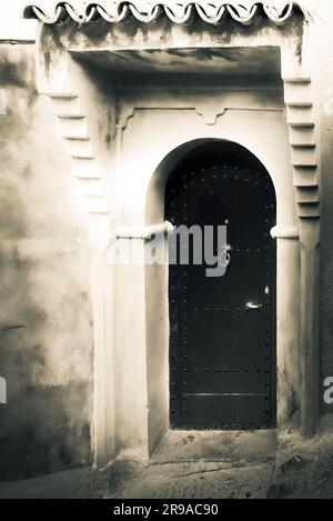 An aged rendering of an An exterior wall and a formidable arched wooden door, framed with architectural details  Chefchaouen, Morocco Stock Photo
