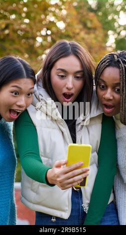 Three multiethnic female ladies amazed watching cellphone, standing outside at city public park. Stock Photo