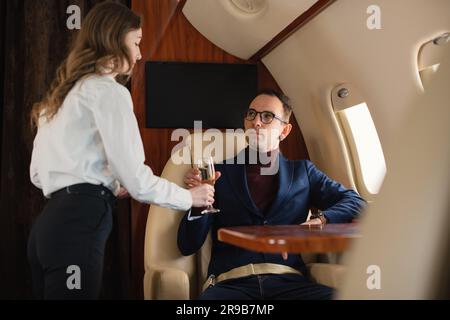 Woman cabin crew serves a glass of champagne to an elegant mid-adult CEO businessman in eyeglasses on a private airplane jet Airline Business Concept Stock Photo