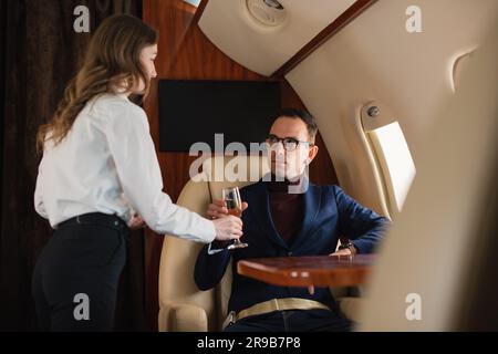 Woman cabin crew serves a glass of champagne to an elegant mid-adult CEO businessman in eyeglasses on a private airplane jet Airline Business Concept Stock Photo