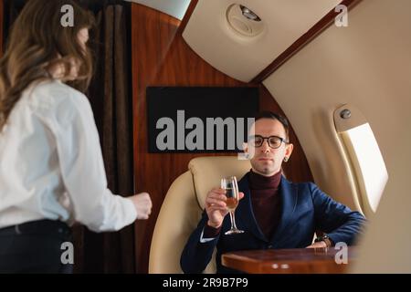 Woman cabin crew serves a glass of champagne to an elegant mid-adult CEO businessman in eyeglasses on a private airplane jet Airline Business Concept Stock Photo