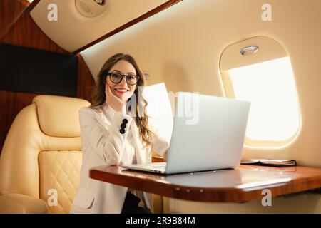 Confident elegant blonde SEO young woman manager in eyeglasses working on the go using a modern laptop while traveling on first class  airplane jet Stock Photo