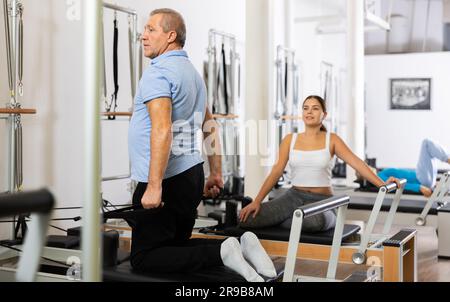 Serious old man in sportswear sitting and doing Pilates exercises for hands on reformer bed equipment in gym Stock Photo