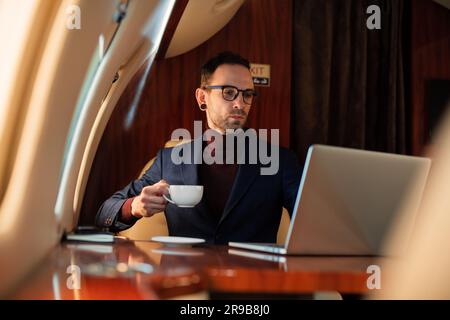 Elegant CEO Young businessman in eyeglasses man sitting inside in first class of private jet drinks coffee after in working on his modern laptop Stock Photo