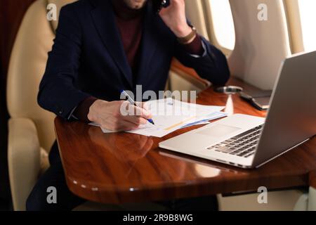 Young elegant CEO businessman in eyeglasses sitting inside a private jet airplane and working in paper currency docs and laptop during the flight Stock Photo