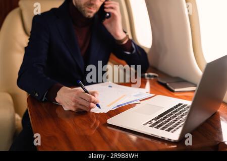 Young elegant CEO businessman in eyeglasses sitting inside a private jet airplane and working in paper currency docs and laptop during the flight Stock Photo