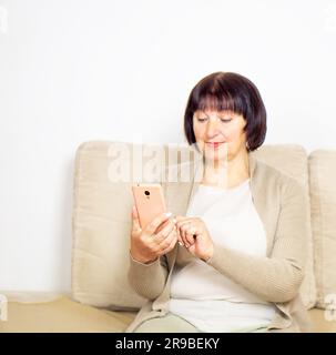 Happy relaxed older woman sitting on comfortable sofa at home and using a mobile phone. Stock Photo