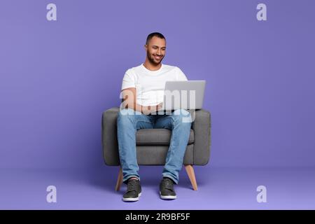 Smiling young man working with laptop in armchair on lilac background Stock Photo