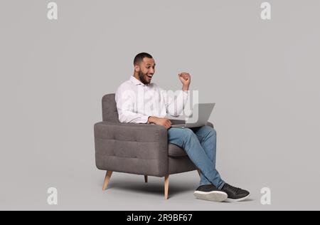 Happy young man with laptop sitting in armchair on grey background Stock Photo