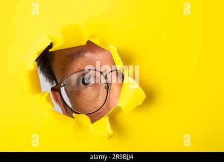 A man wearing glasses peaking through a hole in yellow paper Stock Photo