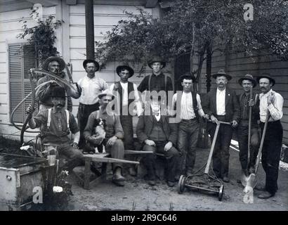 United States:  c. 1900 An older man with his workers and landscaping crew and their tools, including a wheelbarrow, shovels, shears, lawn mower and a scythe. Stock Photo