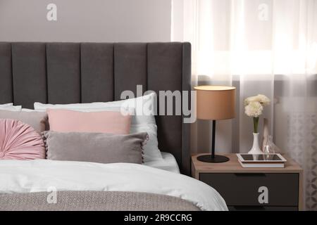 Comfortable bed with cushions, lamp and different decor on bedside table in room. Stylish interior Stock Photo
