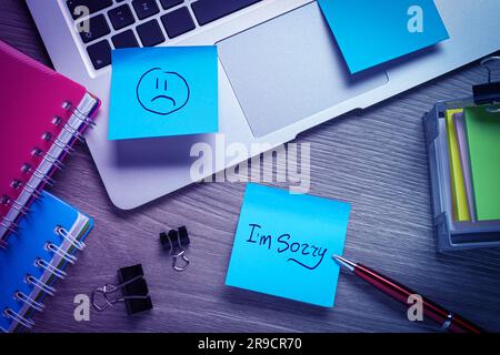 Sticky notes with word Sorry and drawn sad face on workplace. Laptop and stationery on wooden table, flat lay Stock Photo