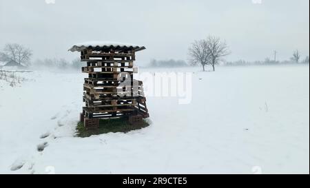 Stack of old used wooden pallets on top of four red building blocks covered with large roof tile and snow left in local field covered with Stock Photo