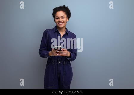 young cheerful positive brunette latin woman with afro hair gathered in a ponytail dressed in a blue stylish denim suit holds a phone in her hands Stock Photo