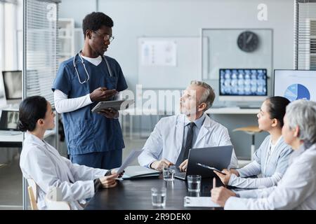 Young confident African American male intern with tablet making speech or report upon medical topic while standing in front of experienced doctor Stock Photo