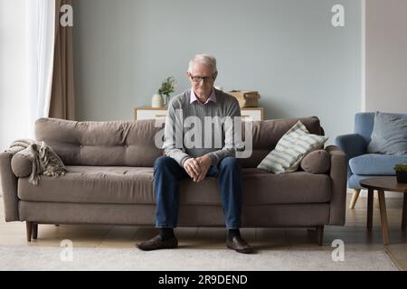Sad thoughtful retired man sits on couch at home alone Stock Photo