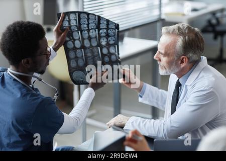 Young African American assistant showing and explaining MRI scan results of brain of patient to mature male radiologist in medical office Stock Photo