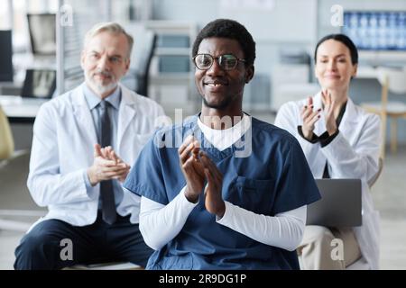 Happy young African American assistant in uniform and two experienced general practitioners on background clapping hands at conference Stock Photo