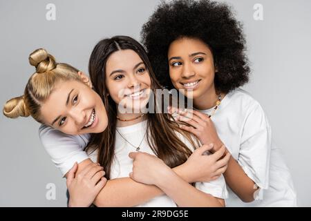 Portrait of cheerful and multiethnic teenagers in white t-shirts hugging and looking away while posing isolated on grey, adolescence girls and generat Stock Photo