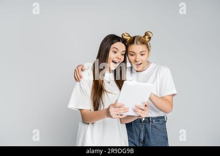 Excited teenage girlfriends in white t-shirts using digital tablet together while standing isolated on grey, teenagers bonding over common interest, f Stock Photo