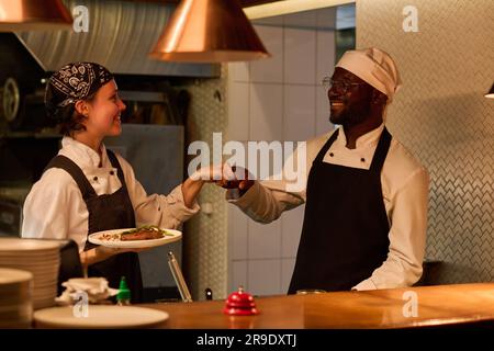 Two happy young intercultural kitchen workers in uniform giving each other punch bump while one of them holding plate with beef steak Stock Photo