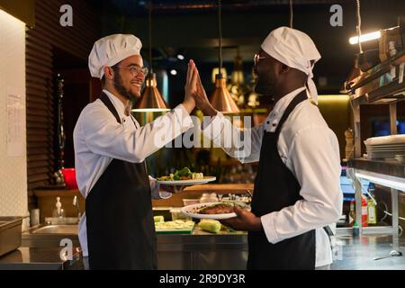 Side view of two happy young intercultural male waiters or cooks in uniform holding plates with food and giving each other high five Stock Photo