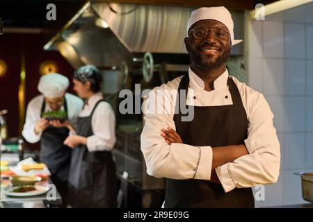 Happy African American male chef in uniform standing in front of camera in restaurant kitchen and keeping his arms crossed by chest Stock Photo