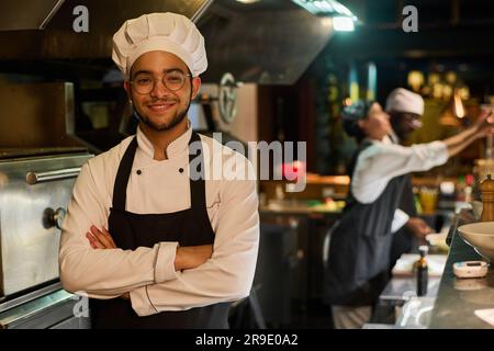 Young smiling cook in uniform keeping his arms crossed by chest and looking at camera while standing against his colleagues in the kitchen Stock Photo
