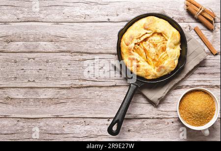 Apple galette in a black pan, brown sugar in a cup and cinnamon sticks, on a wooden table with copy space. Stock Photo