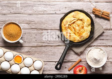Apple galette in a black pan, flour and sugar in cups, cinnamon sticks, a red apple, and some white eggs, on a wooden table. Stock Photo