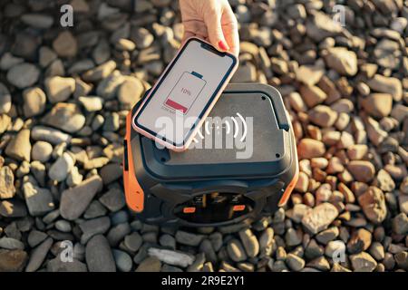 Stylish smartphone with red battery on screen being held by female above portable power station placed on gravel. Innovative renewable energy source providing wireless charging for digital devices. Stock Photo
