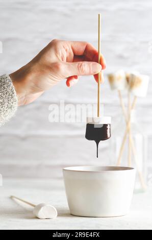 A human hand holding a white marshmallow dripping chocolate into a white cup and some marshmallows in a bottle at the back, on white background. Stock Photo