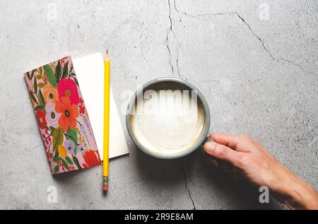 A flowered notebook with a yellow pencil and a right hand holding a cup of latte on a grey stoned background. Stock Photo
