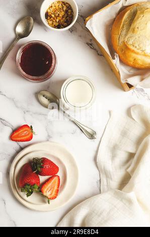 Plain yoghurt, fresh fruits, strawberry jam, a bowl of granola and a loaf of bread with a white napkin on marbled background. Top view. Stock Photo