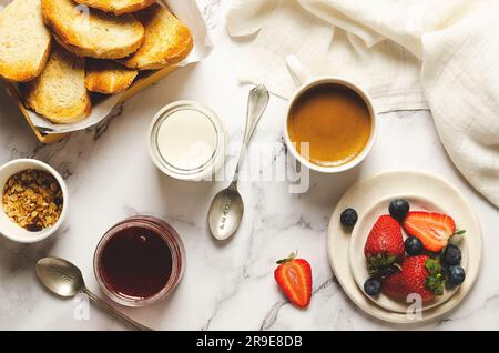 Plain yoghurt, fresh fruits, strawberry jam, a bowl of granola and slices of toast bread with a white napkin on marbled background. Top view. Stock Photo