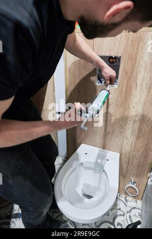Man plumber using plumbing tool applicator gun while installing toilet flush system in apartment under renovation. Male worker applying silicone sealant on corners of toilet flush panel. Stock Photo