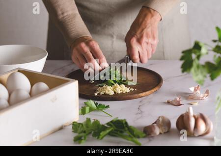 Young woman in a beige apron cuts parsley with a knife. The process of preparing Argentinian milanesas. Stock Photo