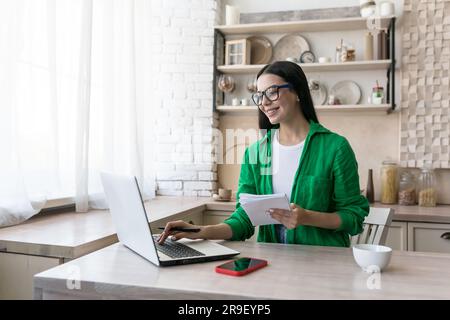 A young freelance businesswoman works remotely from home. Sitting in the kitchen at the laptop and making notes in a notebook. Stock Photo