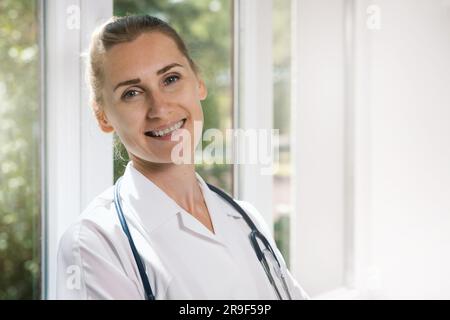 portrait of smiling female doctor with stethoscope standing by the window in hospital hallway. looking at camera. medical staff, medicine student Stock Photo