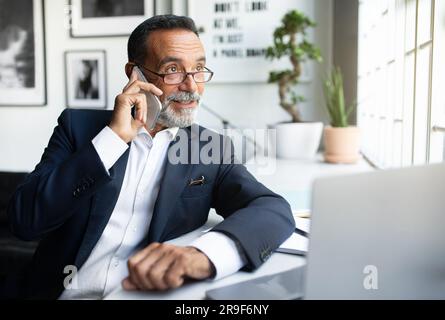 Positive mature european man in suit work at laptop, making phone call in modern office interior Stock Photo