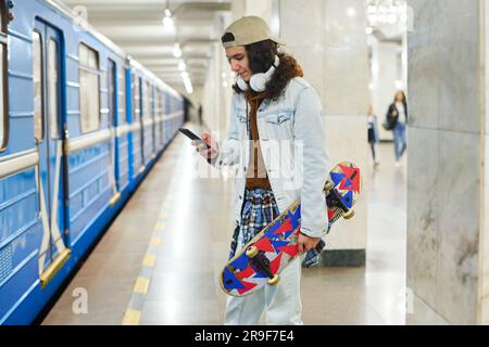 Teenage skateboarder in casual denim attire texting in mobile phone while standing in front of moving blue subway train before catching it Stock Photo