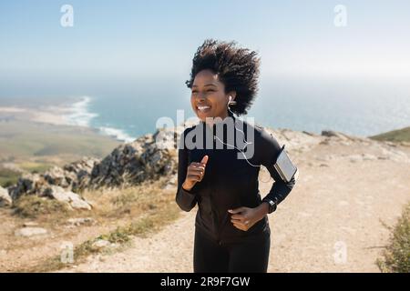Happy young black woman running in morning, training on rocks with ocean view, enjoying workout outdoors Stock Photo