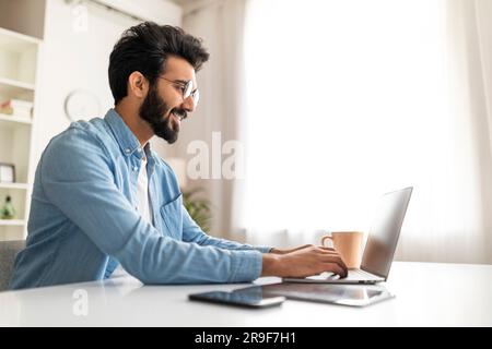 Smiling Indian Male Writer Using Laptop While Working At Home Office Stock Photo