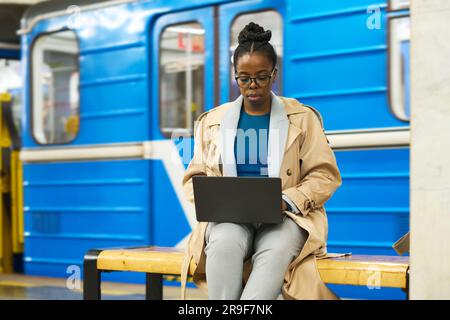 Young African American businesswoman with laptop sitting on bench against blue subway train and organizing work or using video chat Stock Photo