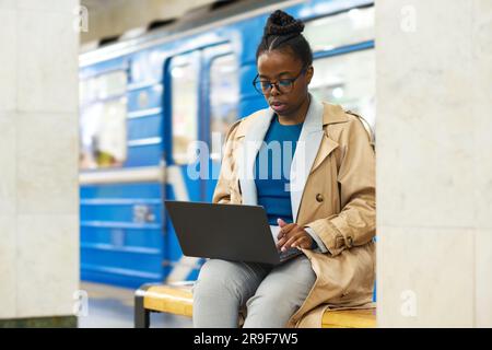 Young serious businesswoman in casualwear networking at subway station while sitting on bench and looking at laptop screen against train Stock Photo