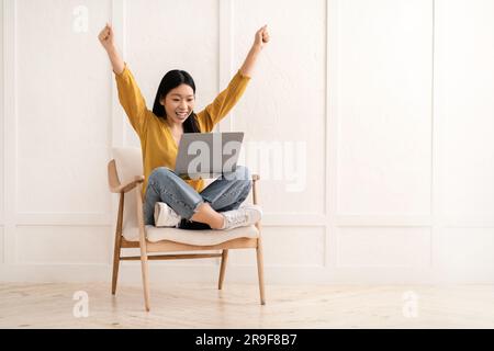 Happy chinese woman sitting in armchair, using computer laptop, gesturing Stock Photo