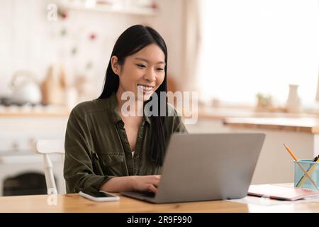 Smiling young asian woman sitting at kitchen, using laptop Stock Photo